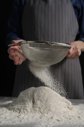 Making bread. Woman sifting flour over dough at table on dark background, closeup