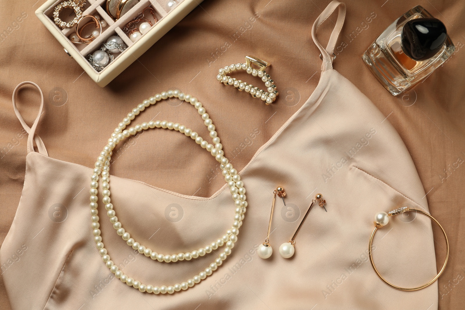 Photo of Stylish jewelry with pearls, luxury perfume and silk dress on beige fabric, flat lay