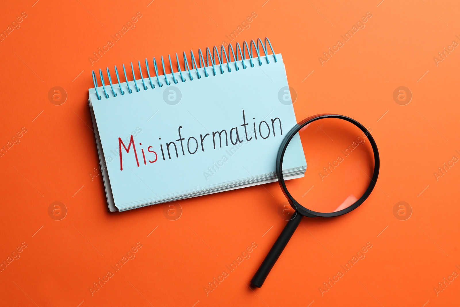 Photo of Notebook with word MISINFORMATION and magnifying glass on orange background, flat lay