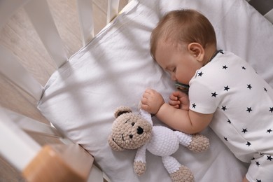 Photo of Adorable little baby with pacifier and toy sleeping in crib indoors, above view