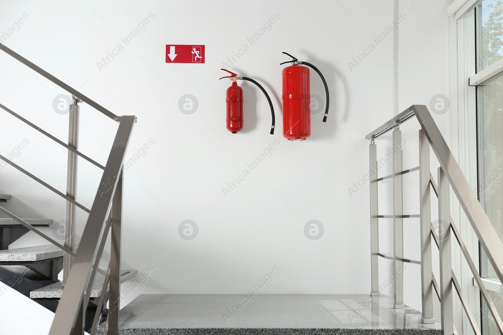 Photo of Fire extinguishers and emergency exit sign on white wall near staircase indoors