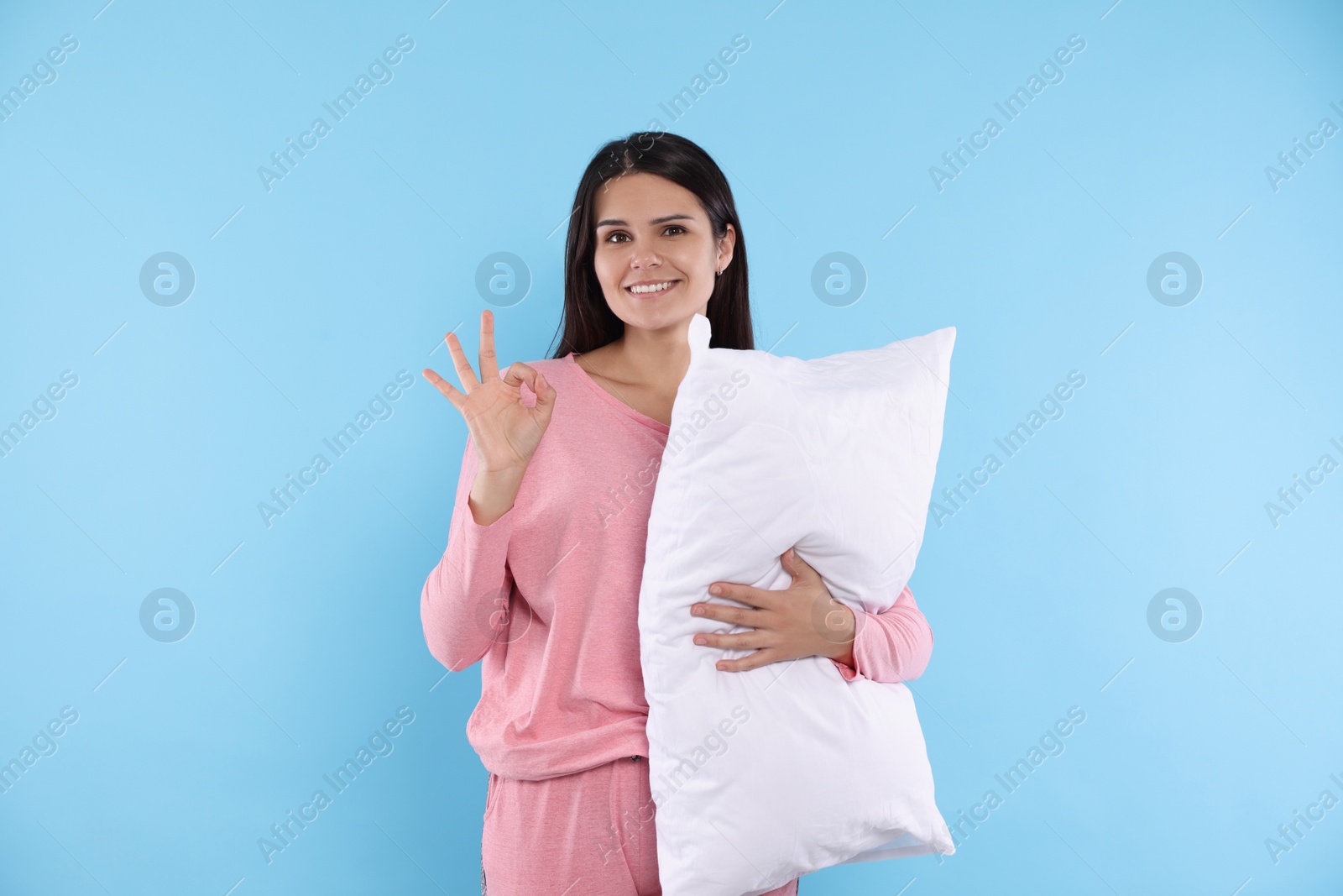 Photo of Happy young woman with soft pillow showing okay gesture on light blue background