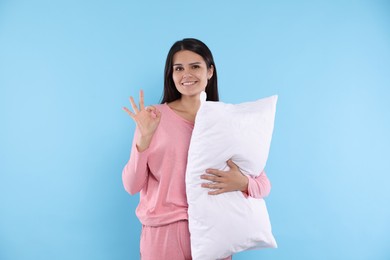 Happy young woman with soft pillow showing okay gesture on light blue background