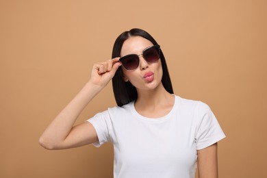 Photo of Beautiful young woman in stylish sunglasses blowing kiss on beige background