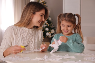 Photo of Happy mother and daughter making paper snowflakes at table near Christmas tree indoors