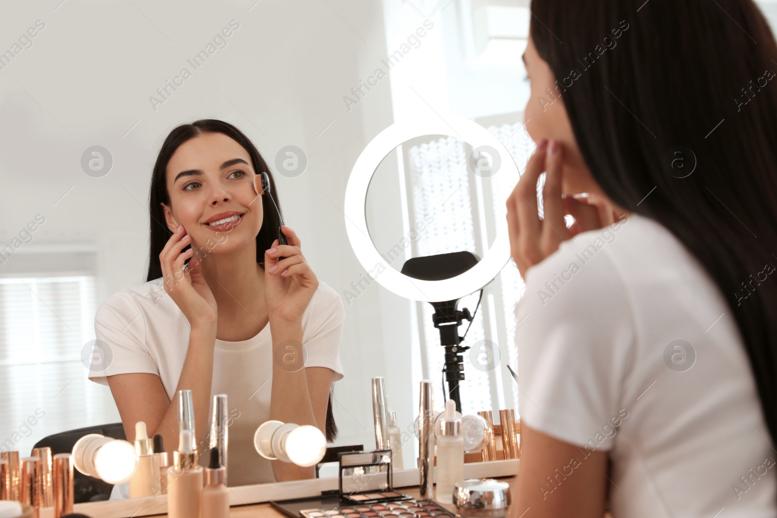 Photo of Beautiful young woman applying makeup at table with mirror and ring lamp