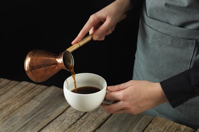 Photo of Turkish coffee. Woman pouring brewed beverage from cezve into cup at wooden table against black background, closeup