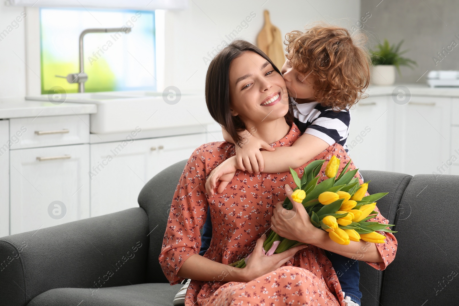 Photo of Little son kissing and congratulating his mom with Mother`s day at home. Woman holding bouquet of yellow tulips