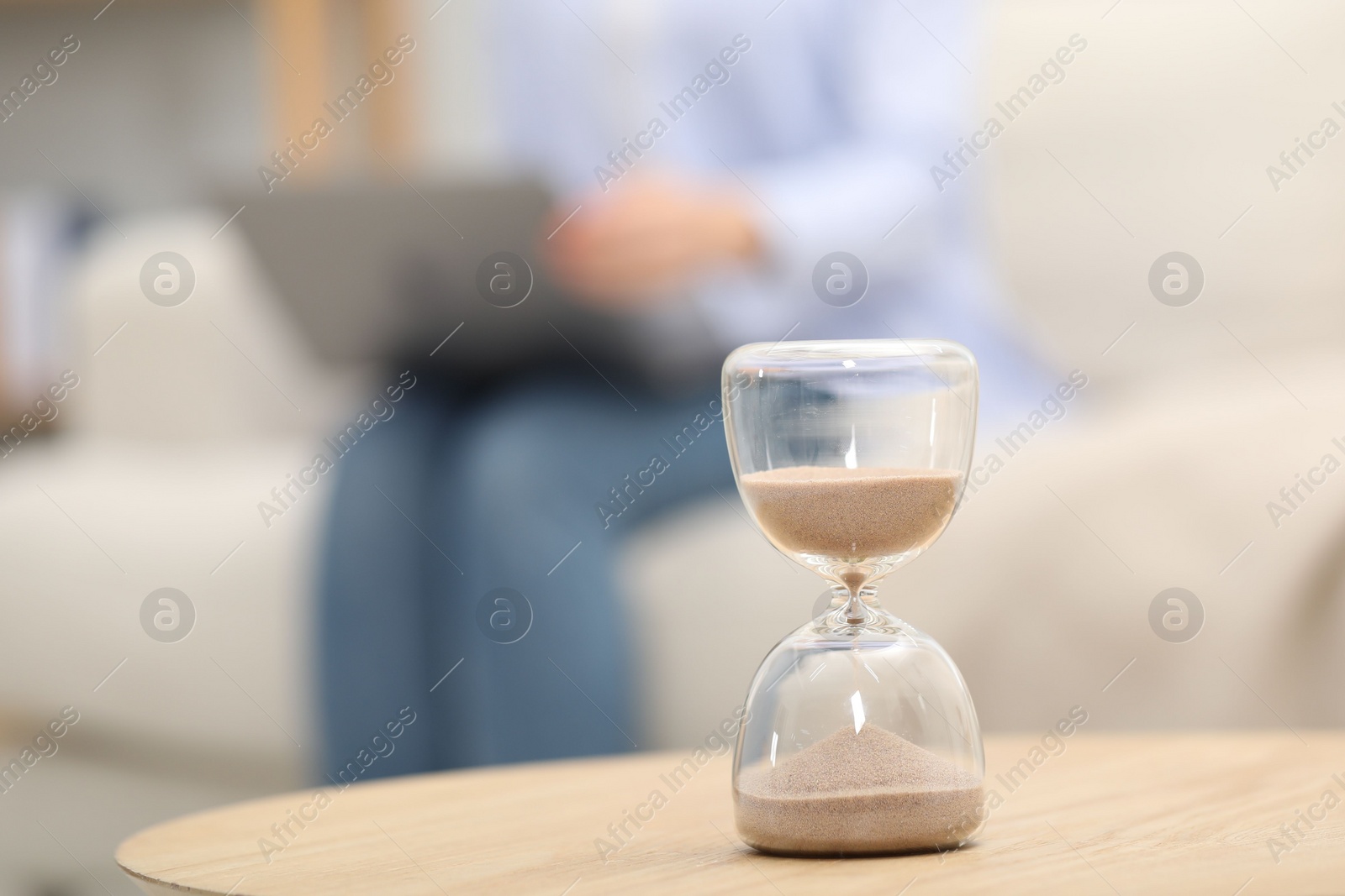 Photo of Hourglass with flowing sand on desk. Woman using laptop indoors, selective focus