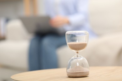 Hourglass with flowing sand on desk. Woman using laptop indoors, selective focus