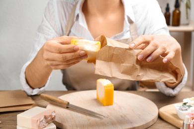 Photo of Woman putting natural handmade soap into paper bag at wooden table, closeup