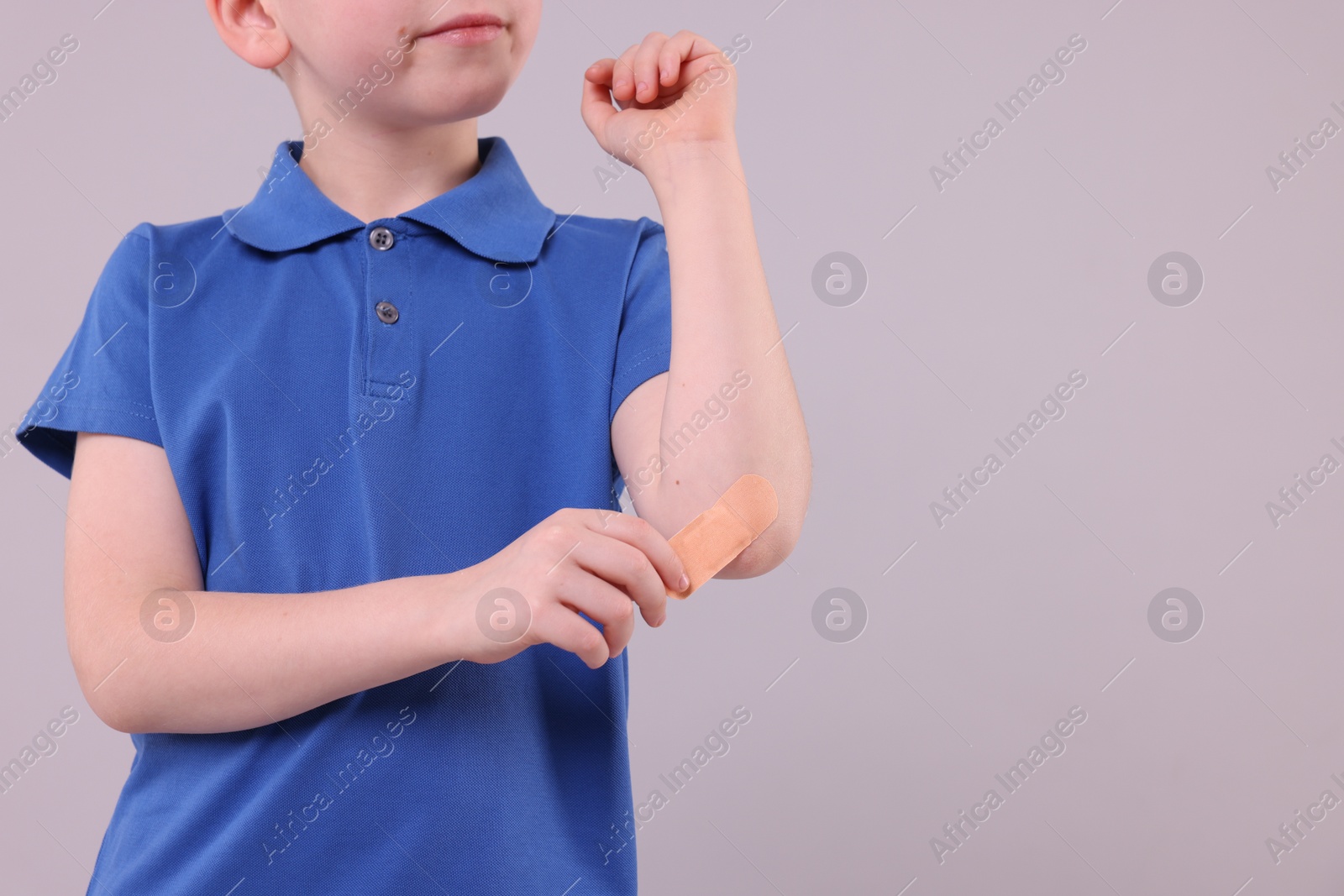 Photo of Little boy putting sticking plaster onto elbow against light grey background, closeup. Space for text