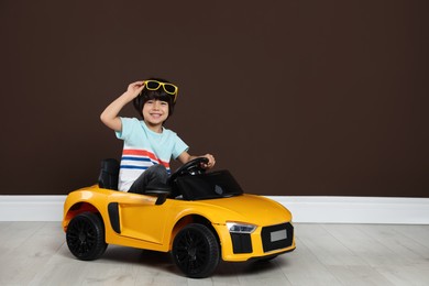 Photo of Cute little boy driving children's electric toy car near brown wall indoors