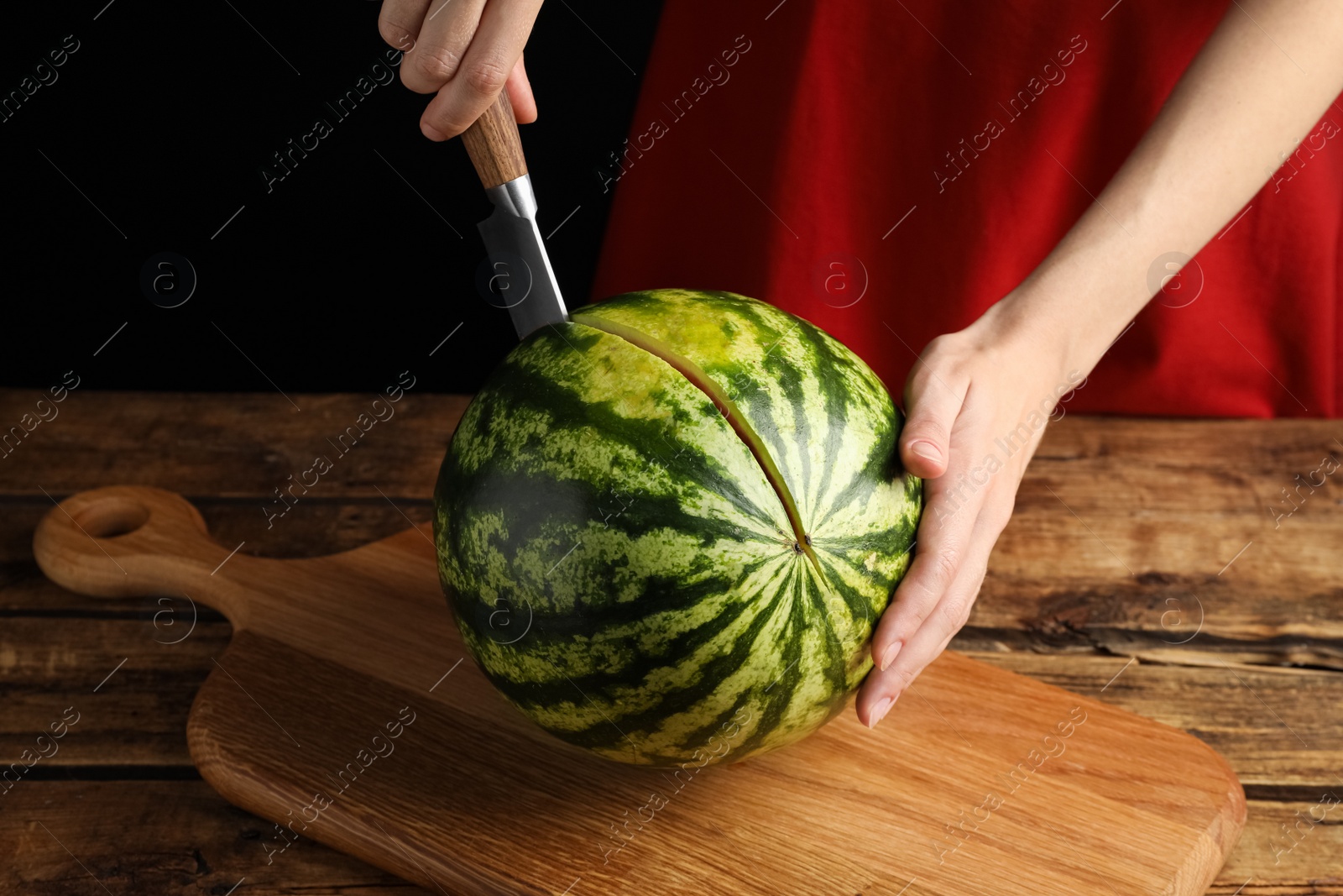 Photo of Woman cutting delicious watermelon at wooden table against black background, closeup