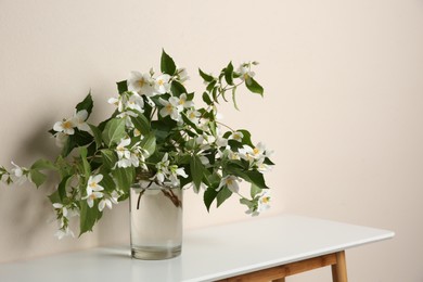 Photo of Bouquet of beautiful jasmine flowers in glass vase on table near beige wall, space for text
