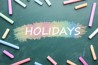 Word HOLIDAYS and pieces of color chalk on greenboard, flat lay