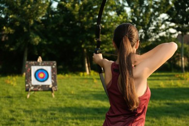 Photo of Woman with bow and arrow aiming at archery target in park, back view