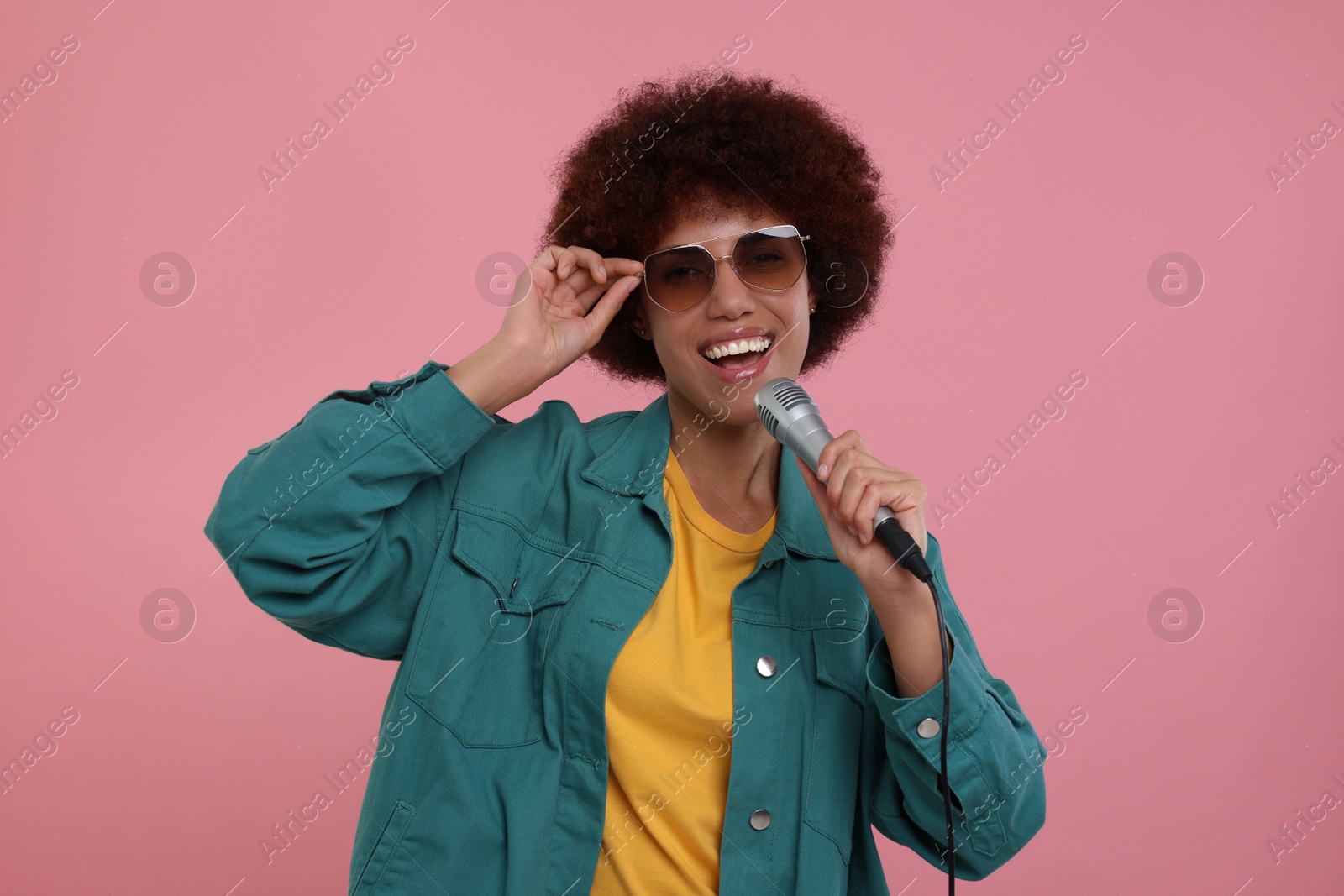 Photo of Curly young woman in sunglasses with microphone singing on pink background