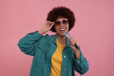 Curly young woman in sunglasses with microphone singing on pink background