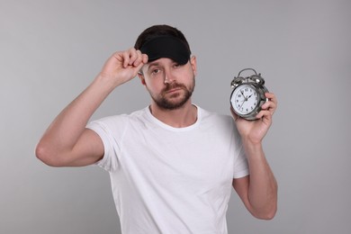 Photo of Sleepy man with alarm clock and mask on grey background. Insomnia problem