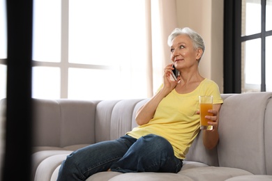 Photo of Mature woman with drink talking on mobile phone at home. Smart aging