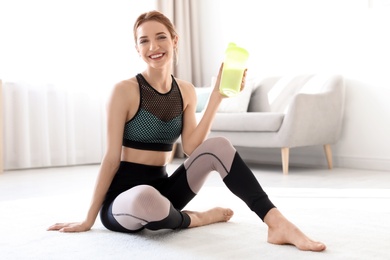 Photo of Young sporty woman with bottle of protein shake sitting on floor at home. Space for text