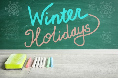 Text Winter Holidays on school greenboard near table with chalk and duster