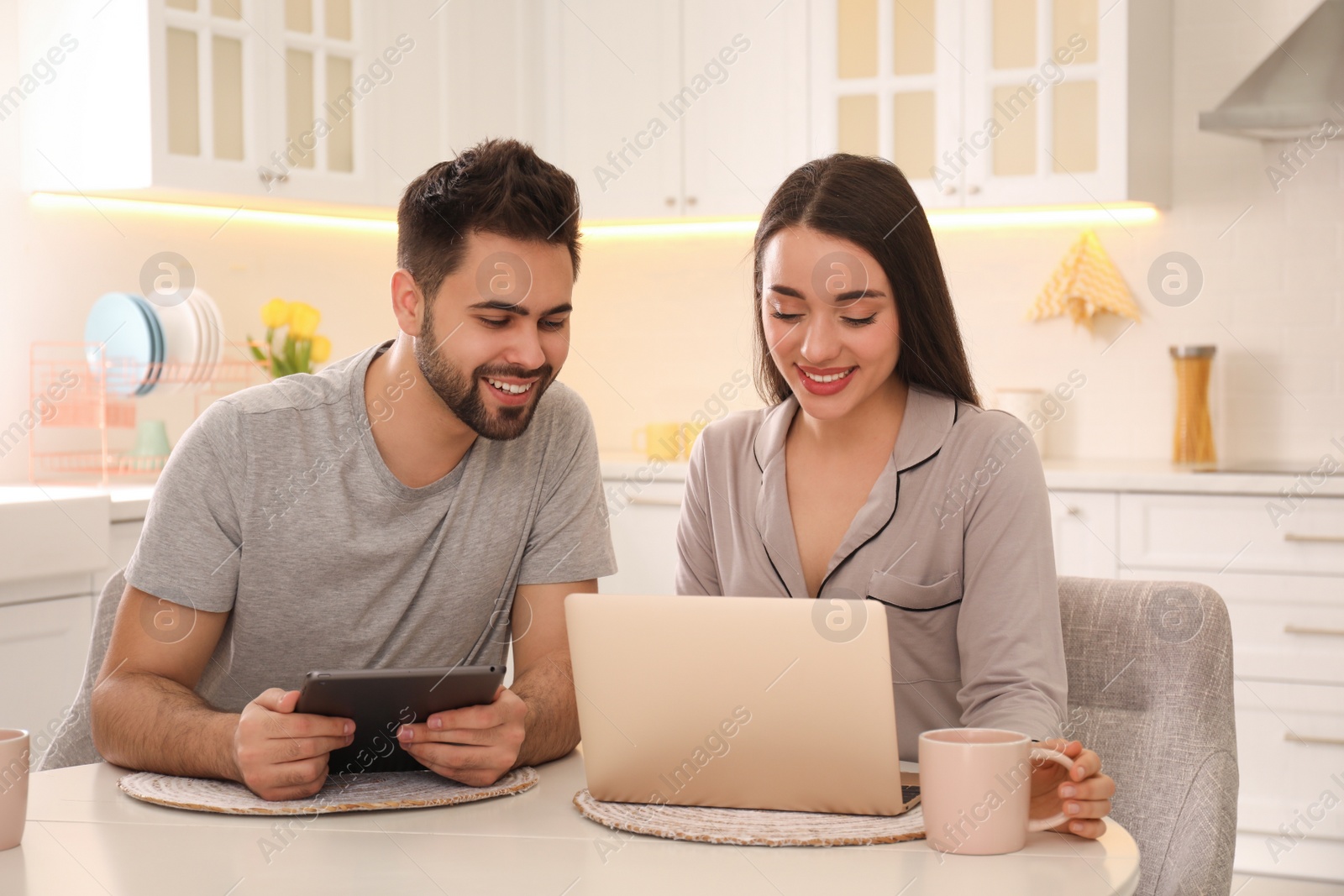 Photo of Happy couple in pajamas using gadgets at kitchen table