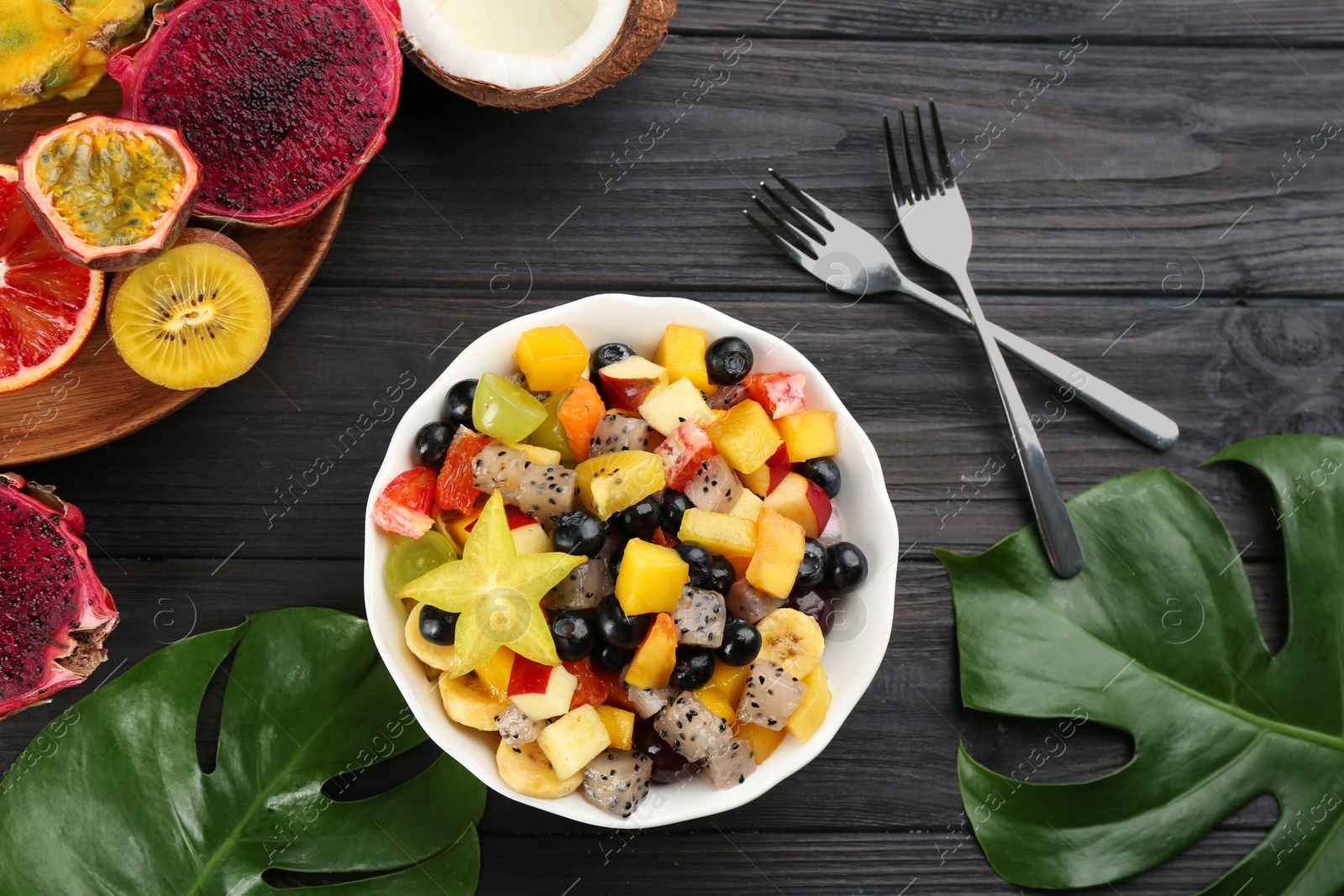 Photo of Delicious exotic fruit salad and ingredients on black wooden table, flat lay