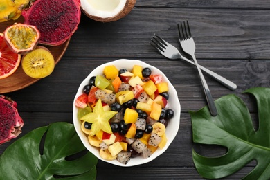 Photo of Delicious exotic fruit salad and ingredients on black wooden table, flat lay