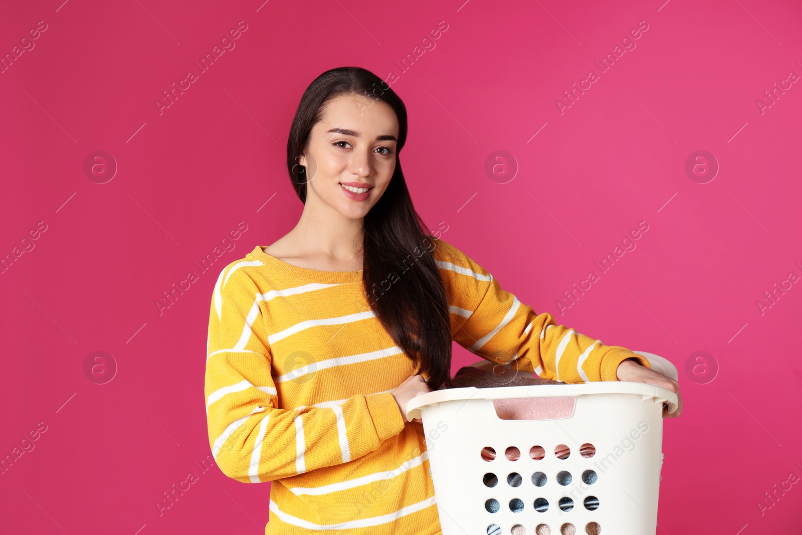 Photo of Happy young woman holding basket with laundry on color background