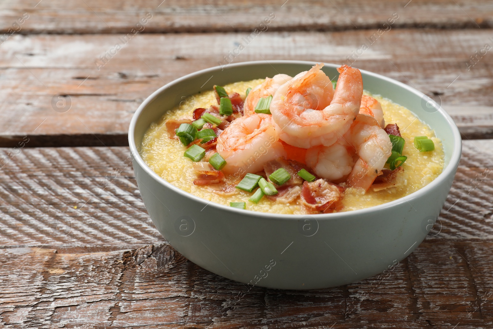 Photo of Fresh tasty shrimps, bacon, grits and green onion in bowl on wooden table, closeup