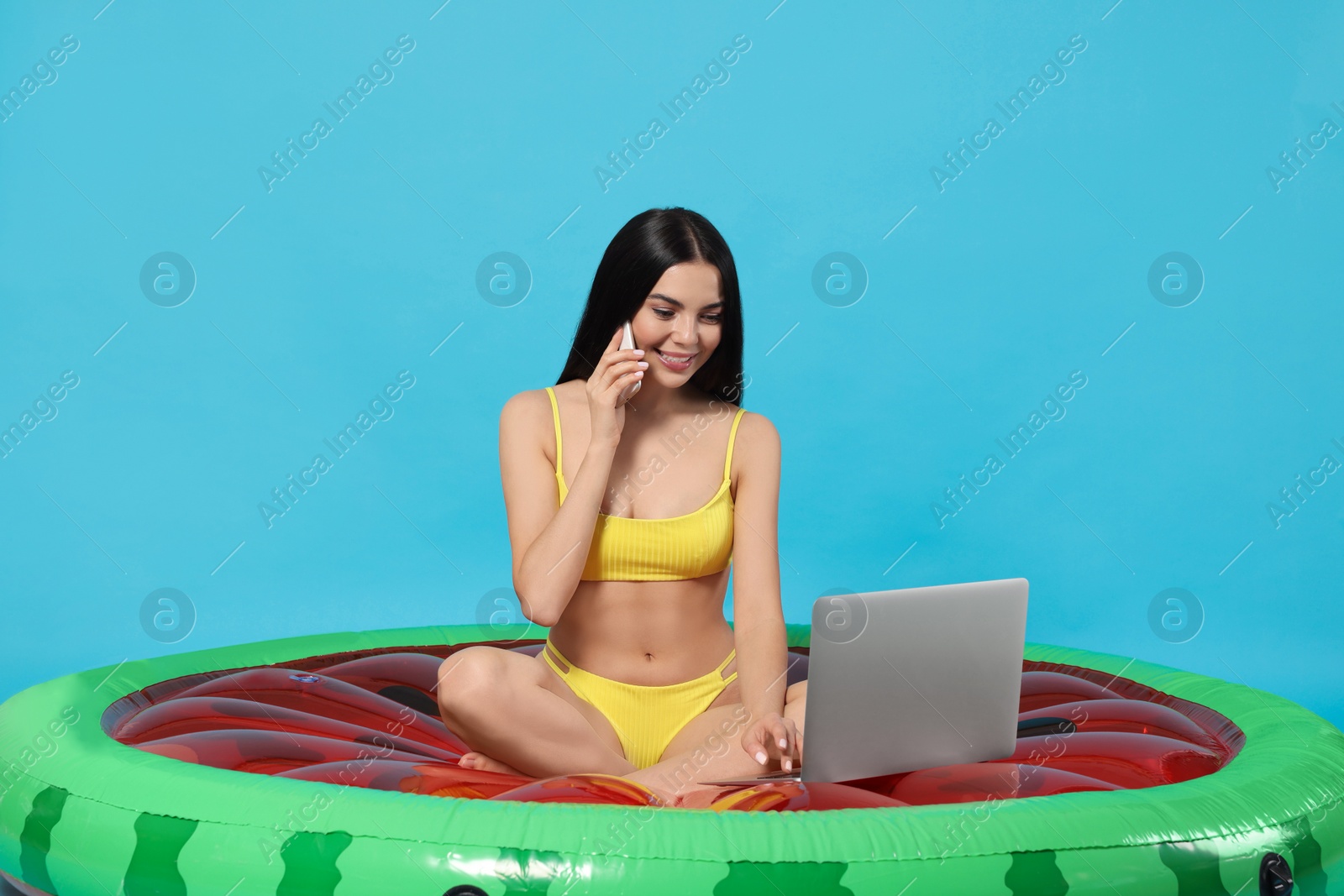 Photo of Young woman with smartphone using laptop while sitting on inflatable mattress against light blue background