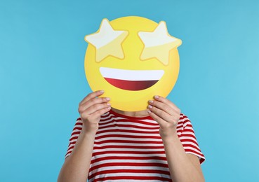 Photo of Woman holding emoticon with stars instead of eyes on light blue background