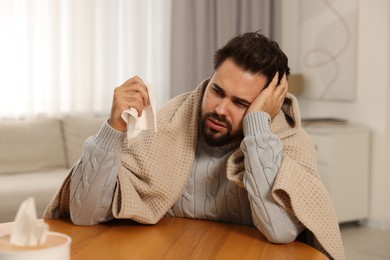 Photo of Sick man wrapped in blanket with tissue at wooden table indoors. Cold symptoms
