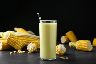 Photo of Tasty fresh corn milk in glass and cobs on grey table against black background