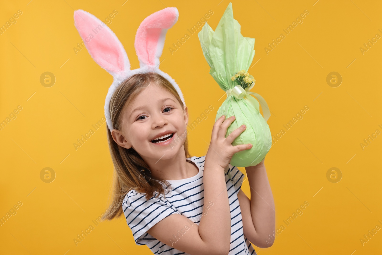 Photo of Easter celebration. Cute girl with bunny ears holding wrapped gift on orange background