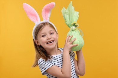 Easter celebration. Cute girl with bunny ears holding wrapped gift on orange background