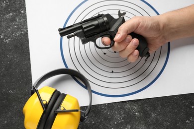 Photo of Man with handgun, shooting target and headphones at dark gray table, top view