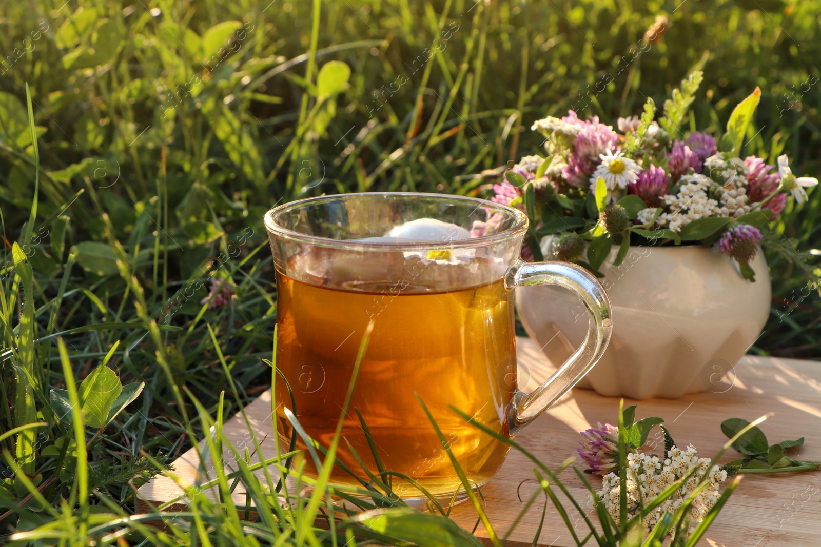Photo of Cup of aromatic herbal tea and ceramic mortar with different wildflowers on green grass outdoors