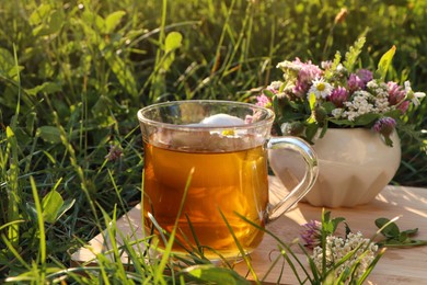 Cup of aromatic herbal tea and ceramic mortar with different wildflowers on green grass outdoors