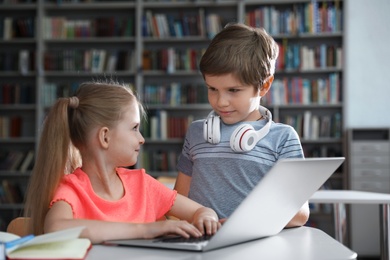 Photo of Little boy with headphones and girl reading book using laptop in library