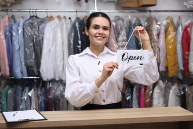 Photo of Dry-cleaning service. Happy worker holding Open sign at counter indoors
