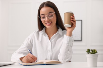 Young woman with cup of coffee writing in notebook at white table indoors