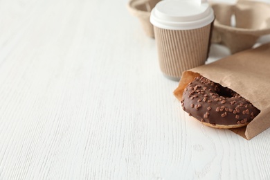 Tasty donut in paper bag and coffee on wooden table. Space for text