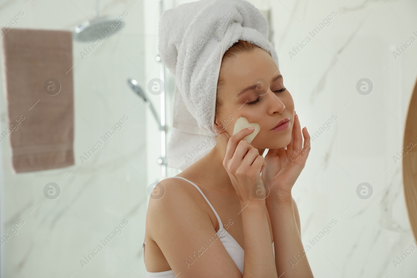 Photo of Beautiful young woman doing facial massage with gua sha tool in bathroom