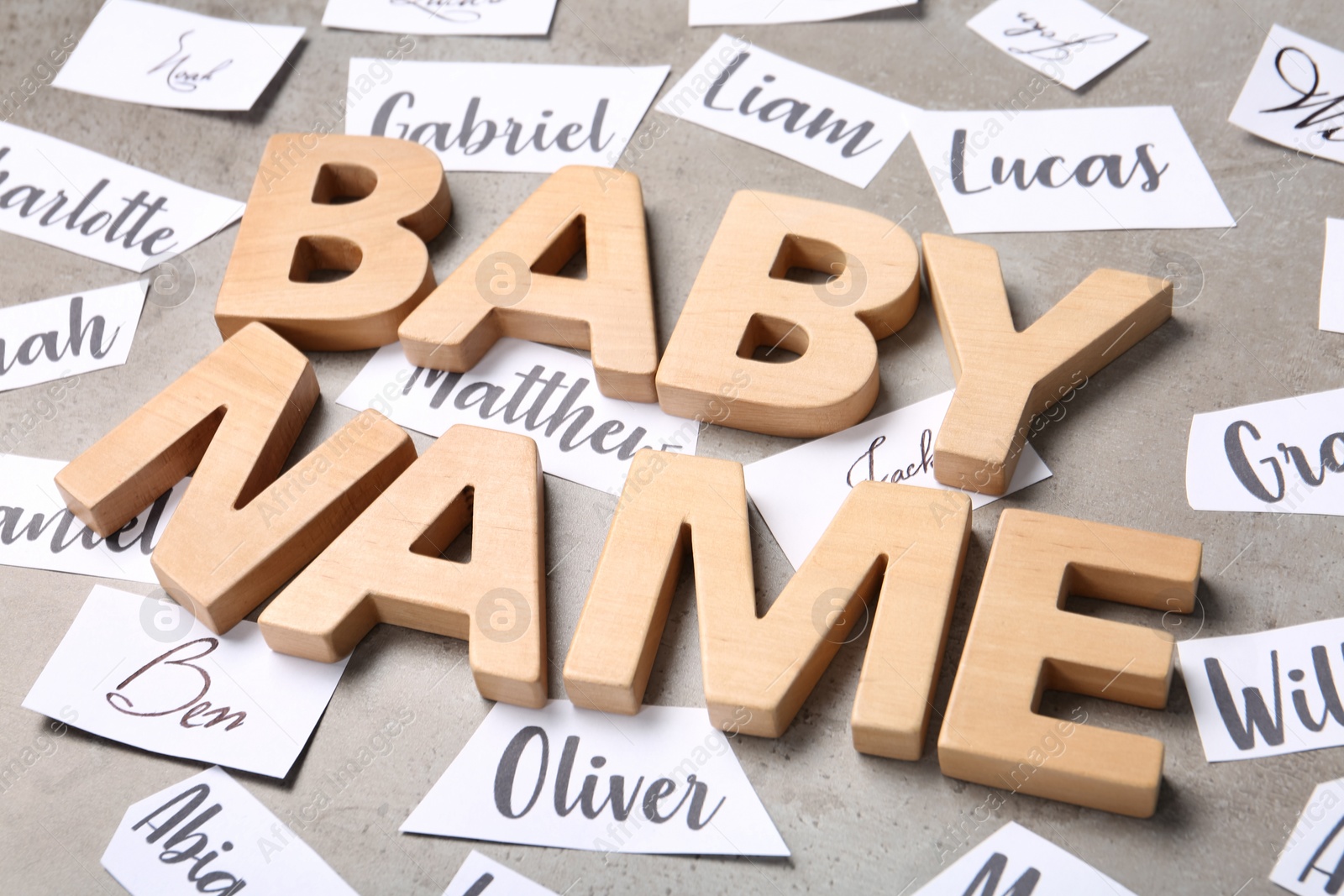 Photo of Paper sheets with different baby names, and wooden letters on grey background, closeup
