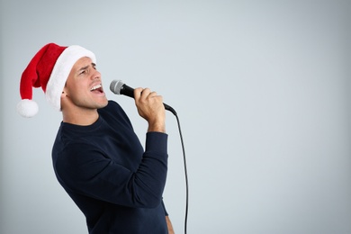 Photo of Emotional man in Santa Claus hat singing with microphone on light grey background, space for text. Christmas music