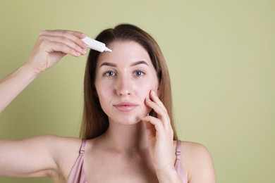 Young woman with acne problem applying cosmetic product onto her skin on olive background