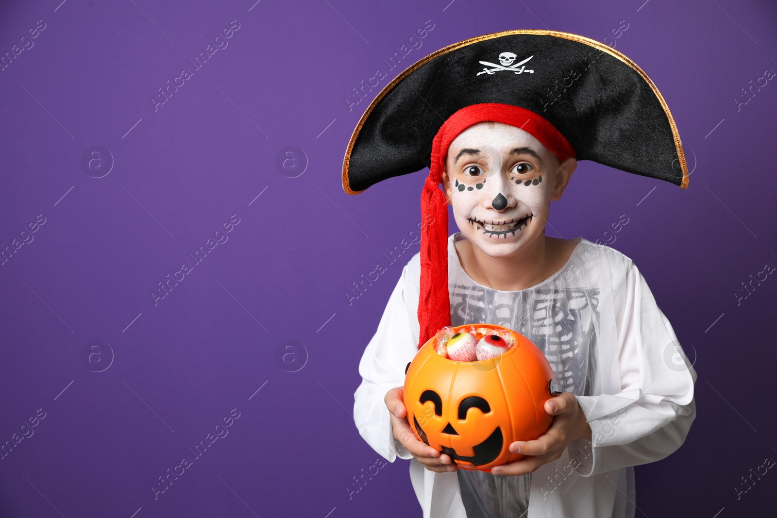 Photo of Cute little boy with pumpkin candy bucket wearing Halloween costume on purple background. Space for text